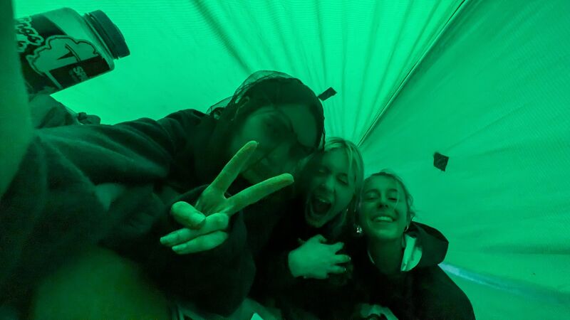 The image shows three people inside a green tent. One person is holding up a peace sign and appears to be taking a selfie with the other two. The lighting is bright green, giving the scene an otherworldly feel. A container is visible in the upper left corner.